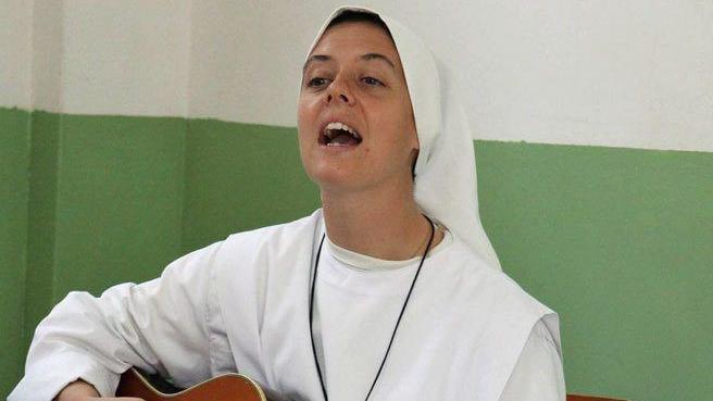 Sr Clare Crockett in white habit, appearing to be singing. she is sat against a green and white wall.