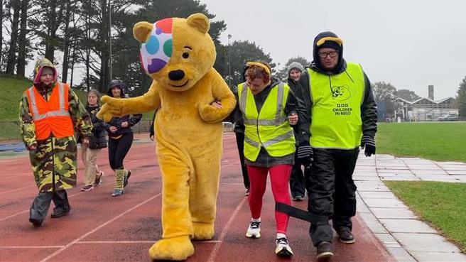 The photo shows radio presenter James Churchfield walking with Pudsey Bear and some guests round Carn Brea running track in Cornwall. They are wearing bright Children in Need branded clothing. They are all looking down at their feet in the rain. 