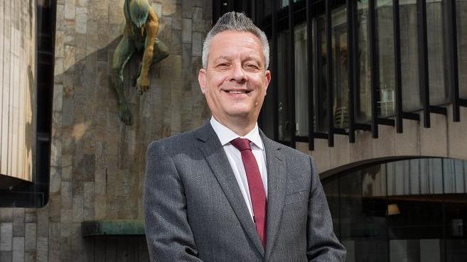 Nick Kemp smiling in front of Newcastle Civic Centre. He's got grey spikey hair and is wearing a dark grey suit and red tie.