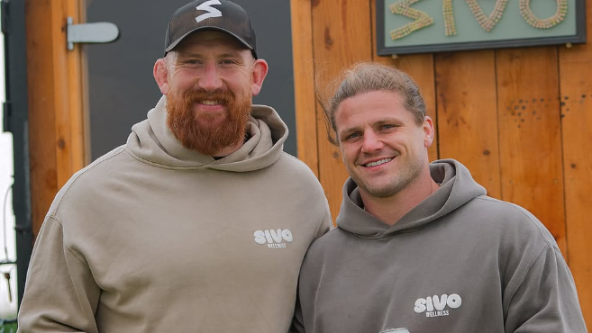 Two Bristol Bears players - Jake Woolmore and Harry Thacker - stand in front of a mobile sauna. Jake has a full beard and wears a baseball cap. Harry has his hair tied back. They are both wearing hoodies and smiling.