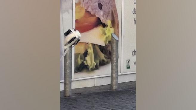 A black square box strapped to a metal bollard with white tape. It is outside a Nisa store.
