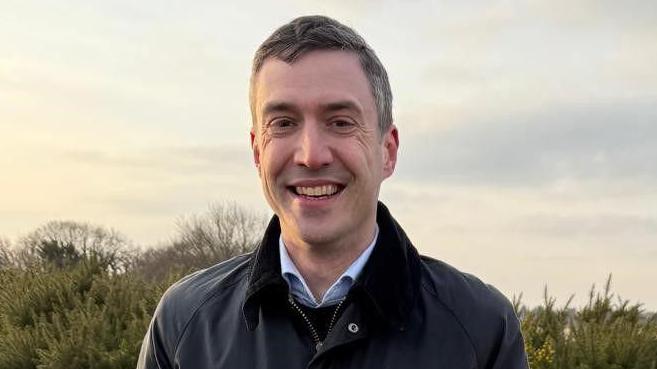 Adrian Ramsey smiling at the camera against the background of some shrubbery. He is wearing a grey waterproof coat and has dark hair.