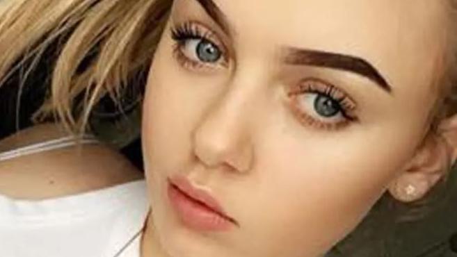 A young white girl with thick brown manicured eyebrows, blonde hair and blue eyes, stares at the camera