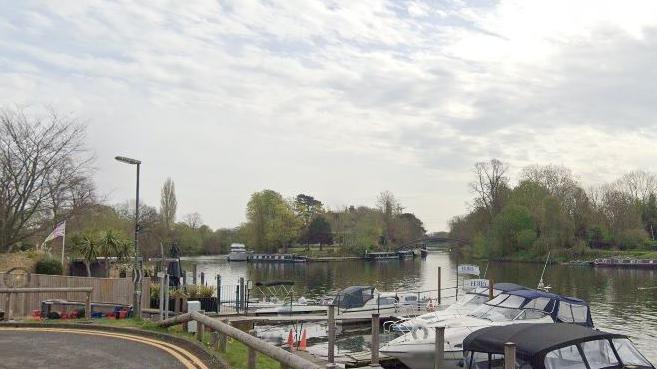 A view of an island in the Thames, seen from a nearby road. There are boats in the foreground and around the island there are boats moored. There are trees around, some in leaf and others are bare while there are white clouds in the sky.