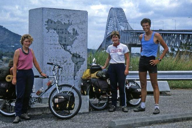The three friends are standing next to a concrete block with an illustration of America on it. They have their bikes and there is a large bridge in the background.
