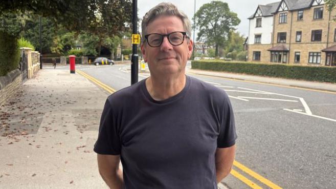 A man in a navy blue t-shirt wearing glasses stands on a pavement in an area of Leeds near Roundhay Park.
