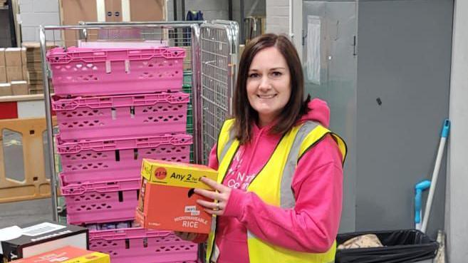 A lady in a high visibility jacket holding a box of food with pink crates in the background. She is wearing a pink hoodie underneath and is holding a box of rice packets.