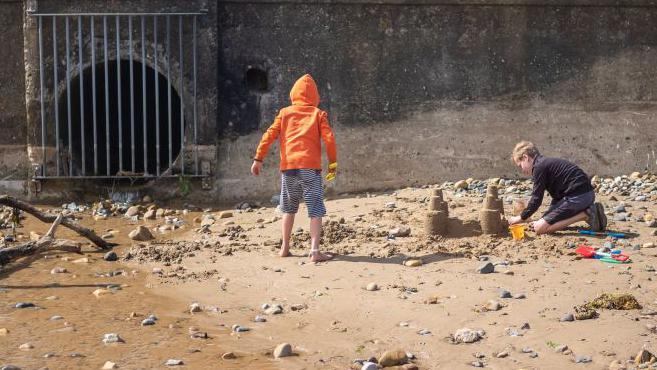 Two children build sand castles next to a storm drain outlet.