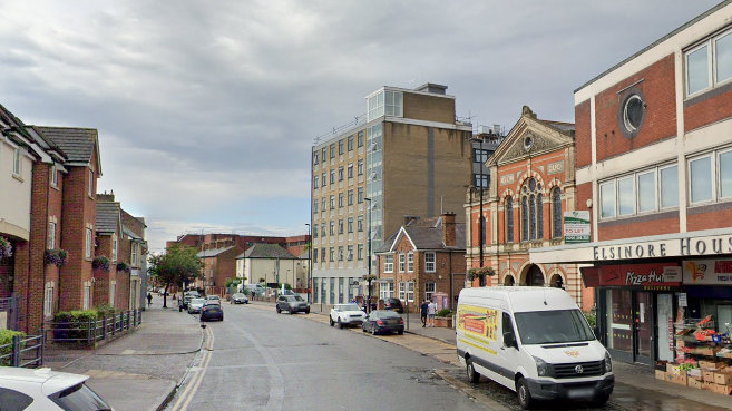 A general view of Buckingham Street in Aylesbury. Vehicles are parked either side of the road while buildings including flats and a parade of shops line either side of the road.