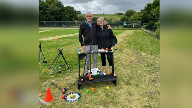 Steve Wilson and Janet Wylie, a white couple with their arms around each other, stand on the park golf course with mallets and colourful balls at their feet