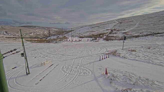 A snowy scene surrounded by hills covered in a light dusting of snow. The image is taken from a webcam mounted on a pole above the ground, with a snow-covered round in the middle of the shot, covered in tyre tracks. 