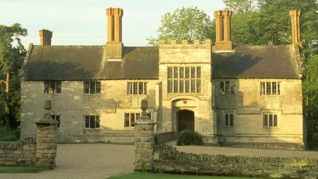 The stone house stands in full view with a stone wall and entrance surrounding it. Three chimneys can be seen with a large door entrance.