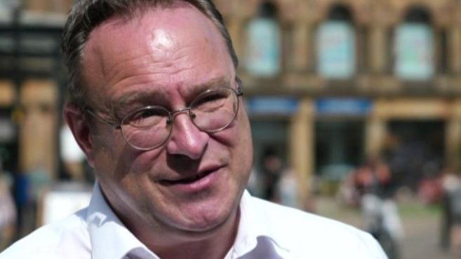 A man stood outside in the daylight with round glasses and wearing a white shirt.