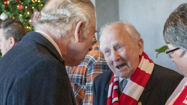 Arthur Massey wearing a red and white scarf, chats with King Charles at Wrexham's football ground