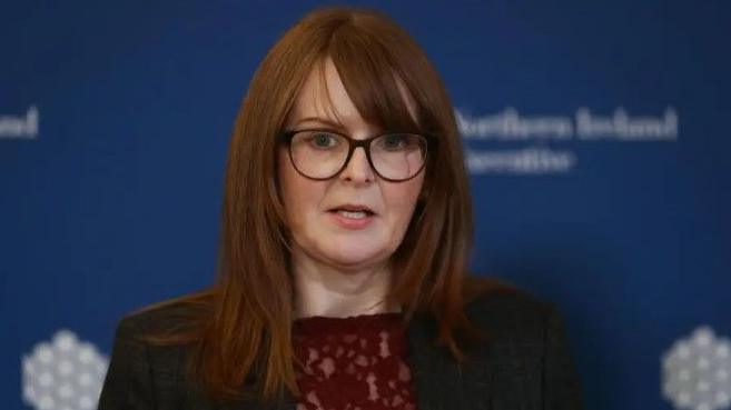 Caoimhe Archibald wearing a dark coloured suit with a blue background behind her.