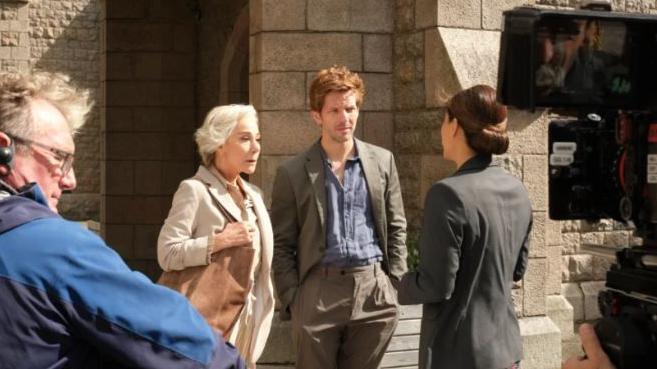 Damien Molony and Zoe Wanamaker in character during filming of the rebooted Bergerac series in Jersey. A camera crew is watching on as Damien and Zoe talk to another actor.