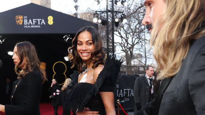 Zoe Saldana, wearing a black dress, smiles on the red carpet at the Bafta film awards as she holds the hand of her husband Marco Perego-Saldana