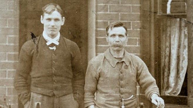 A young man, with a middle parting hair cut and wearing a cardigan, is standing next to his father who has a very thick moustache. He is wearing a jacket, with a pocket watch extending from it. 