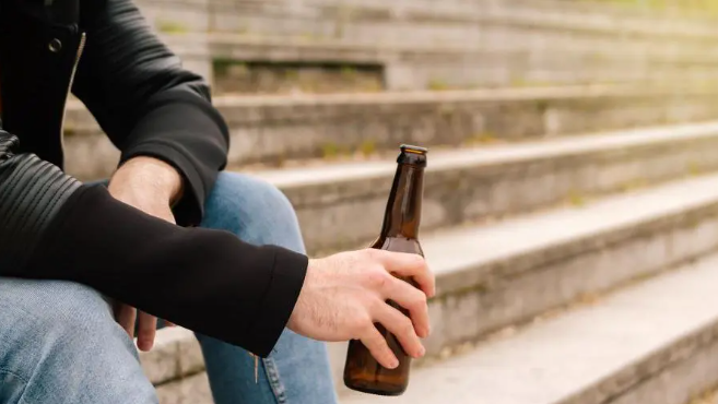 A person in a black hoodie and blue jeans is sat on a step while holding a brown beer bottle