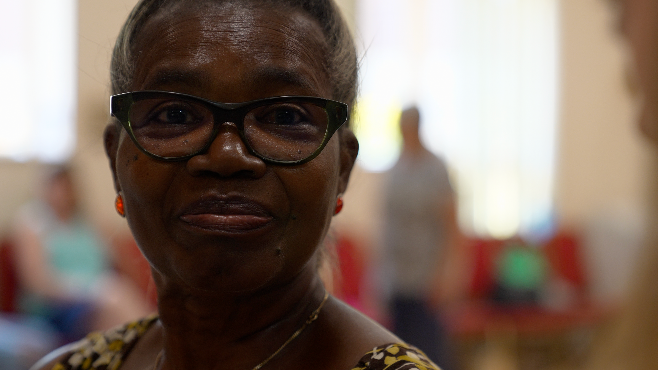 An older woman with greying hair named Vadney, wearing black-rimmed glasses and red earrings