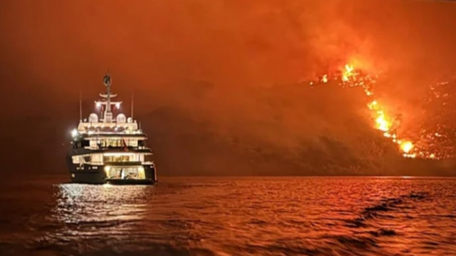 A photo of a luxury yacht on the water, with a raging fire on the land in the distance