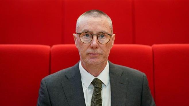 NUH chief executive Anthony May is pictured looking at the camera. He is stood in front a group of red seats and wearing a white shirt, dark grey blazer and olive tie