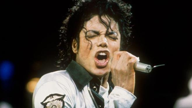 US singer Michael Jackson performing at a 1998 concert in London. He is wearing a white navy jacket with a white collar and singing into a microphone with his eyes closed