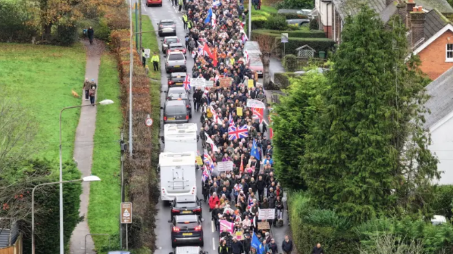 A drone shot of a protest held in Crowborough, East Sussex on 16 November over plans to house asylum seekers at an army site in the town. Hundreds of people can be seen walking on the streets with flags and banners. Cars are driving alongside the walkers.