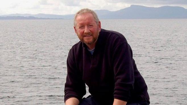 Alan Beattie Herriot is sitting on a rock with the sea behind him. He has short hair and a beard.