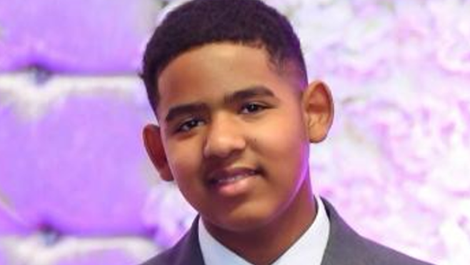 A young man with short black hair is dressed in a suit and white collared shirt. He is looking at the camera, and standing in front of a purple background.