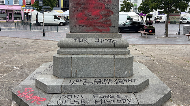 The David Lloyd George statue with black graffiti with the words 'for shame', 'don't commemorate a coloniser' and 'don't forget Welsh history' daubed on the plinth. The town high street is in the background with traffic and shops visible.