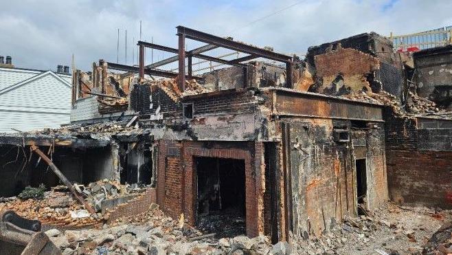 The charred ruins of the burnt out pub. Some of the brick walls of the basement level are still standing and some blackened steel girders remain on the upper level nearest the road but the building is mostly collapsed or demolished.