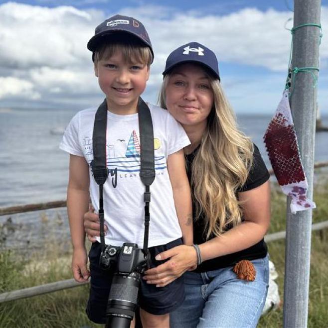 Young boy with camera and his mum, both smiling, with sea in background.