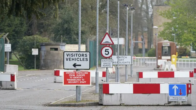 The image shows the entrance to a former military facility, viewed from a road leading up to a checkpoint area. The entrance is controlled with barriers and multiple warning signs. In the front-left of the image, a rectangular white sign with black text reads: VISITORS VEHICLES with a left-pointing arrow. Below it, a red sign with white text states: DRIVERS TRAVELLING FROM EUROPE TO IDENTIFY THEMSELVES TO SECURITY STAFF