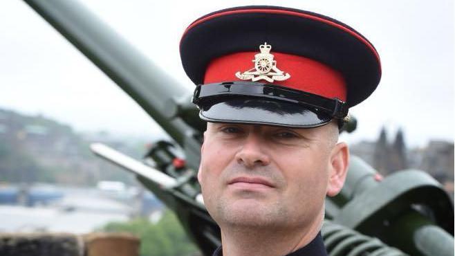 Ryan Brown pictured when he was in the army he is looking into the camera with short white hair wearing a black and red military hat with an emblem in gold. He is standing in front of large military weapons. 