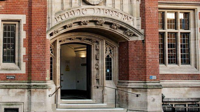 The outside Ipswich County Library. The red brick building has an arch over the entrance with the words 'public library' on this arch. Steps lined with black rails lead up to the entrance.