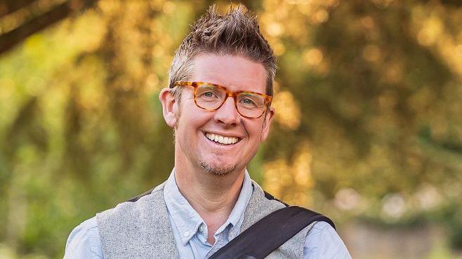 Barnaby Aldrick smiling, wearing brown speckled glasses and wearing a blue top with a grey waistcoat and a black strap on one shoulder. He is standing outside, with a blurred background of trees. His brown hair is gelled in a spiky style and he has stubble on his chin.