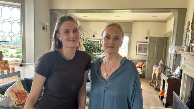 A recent image of Imke, who is now in her 20s, and her mother Caroline, who are looking to camera standing in a large living room with a bay window to the left and a fireplace, desk and rocking horse to the right.
