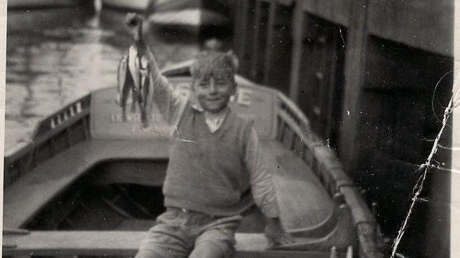 A black and white photograph of a little boy sitting in a fishing boat holding up whiting.