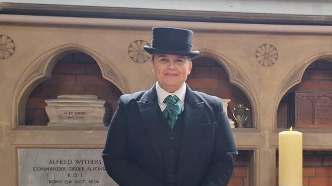 Ms Whymark smiles at the camera while she stands inside a church. She wears a smart black coat, waistcoat, white shirt, green tie and black hat.