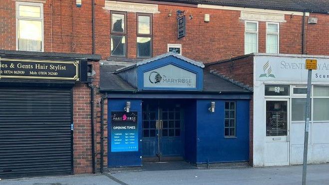 A small bar on a high street, it is painted blue and has signs up stating 'The Marie Rose'. 