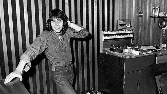 Black and white image of Mark in the 70s in a recording studio. He has shoulder-length hair and is smiling at the camera.