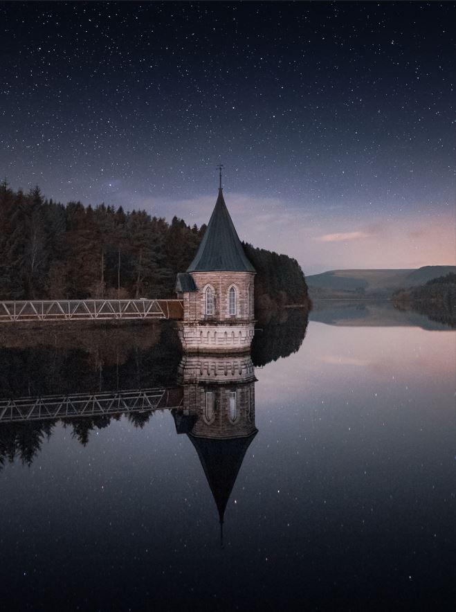 Pontsticill Reservoir in Powys with the valve tower and bridge over the water pictured against the backdrop of starry skies. The tower and bridge are reflected perfectly in the water below and there are trees and mountains in the background.