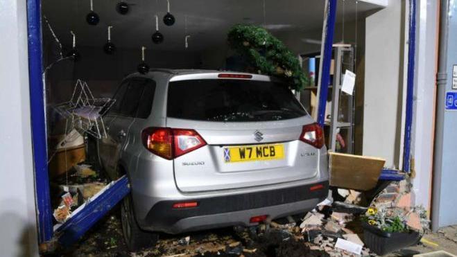 A rear view of a silver car which has smashed through an office window leaving debris all around