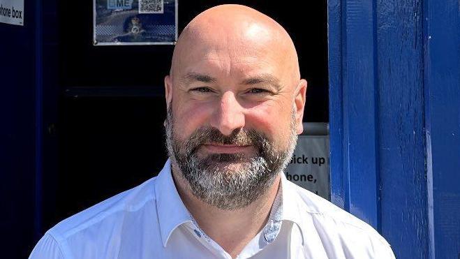 Marc wearing a white shirt and smiling as he sits in front of a blue-coloured small booth