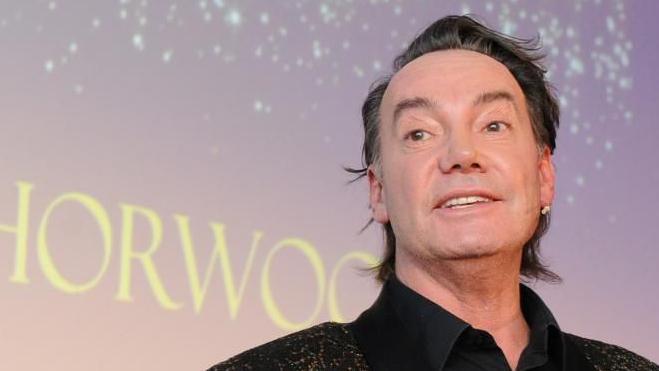 Craig Revel Horwood. He has dark hair, is wearing a stud in one ear, a gold spangled jacket over a black shirt, open at the neck. He is in the middle of speaking. Behind him is a screen. 