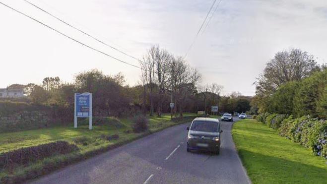 Google Street View of the A392 at White Acres near Newquay. Three vehicles are on the road heading towards the camera. A blue sign for White Acres Holiday Park is on a grass verge on the left-hand side. Trees and a stone wall line the road.