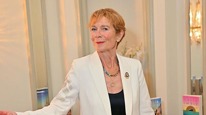 Actress Celia Imrie stood in front of a pile of her books. She is wearing a white jacket and black shirt. Her first arm is slightly raised and she is pointing her index finger.