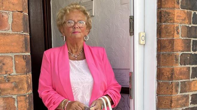 Margaret Scaldwell is standing in the doorway of her home. She is wearing a bright pink blazer and white patterned top.