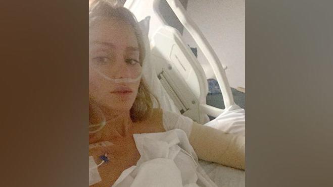 Charlene Morrissey lies in a hospital bed. She has blonde hair. She is hooked up to various machines and wrapped in bandages.